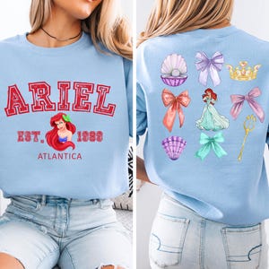 Disney Ariel sweatshirt in pastel blue with bold red varsity lettering on the front, reading 'Ariel Est. 1989 Atlantica.' The back design includes princess elements like seashells, bows, a golden trident, and an elegant illustration of