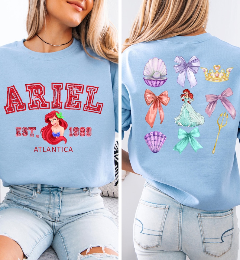Disney Ariel sweatshirt in pastel blue with bold red varsity lettering on the front, reading 'Ariel Est. 1989 Atlantica.' The back design includes princess elements like seashells, bows, a golden trident, and an elegant illustration of