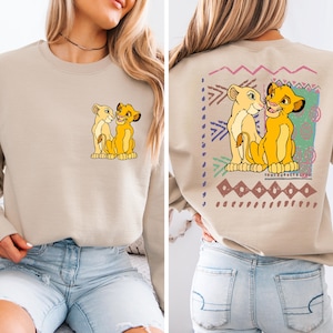 May include: A beige sweatshirt with a graphic print of Simba and Nala from The Lion King. The print features a colorful tribal pattern in the background.