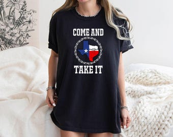 May include: Black t-shirt with the words "COME AND TAKE IT" in white block letters. The Texas state flag is in the center, surrounded by a barbed wire circle. The shirt is a casual style.