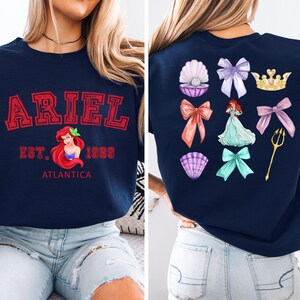 Disney Ariel sweatshirt in pastel blue with bold red varsity lettering on the front, reading 'Ariel Est. 1989 Atlantica.' The back design includes princess elements like seashells, bows, a golden trident, and an elegant illustration of