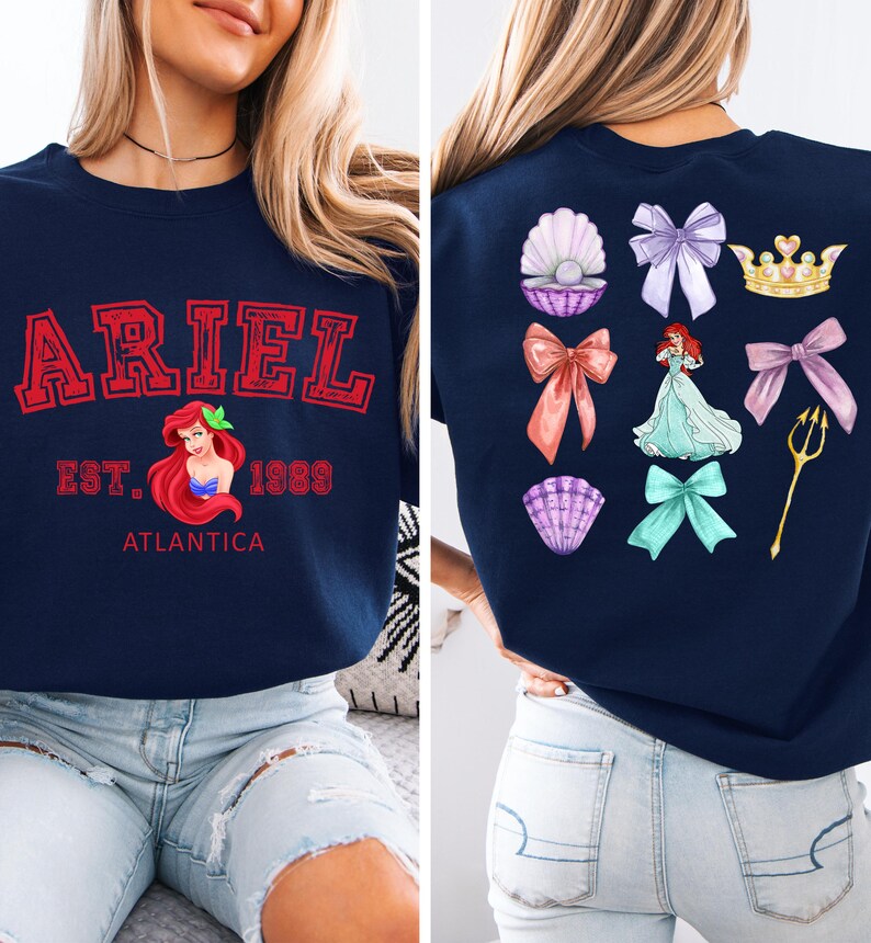 Disney Ariel sweatshirt in pastel blue with bold red varsity lettering on the front, reading 'Ariel Est. 1989 Atlantica.' The back design includes princess elements like seashells, bows, a golden trident, and an elegant illustration of