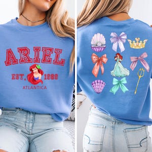 Disney Ariel sweatshirt in pastel blue with bold red varsity lettering on the front, reading 'Ariel Est. 1989 Atlantica.' The back design includes princess elements like seashells, bows, a golden trident, and an elegant illustration of