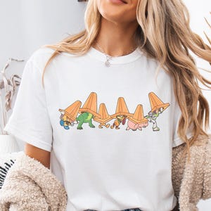 May include: White t-shirt with a graphic of Toy Story characters wearing traffic cones on their heads. The characters include Woody, Buzz Lightyear, Rex, Hamm, and Slinky Dog.