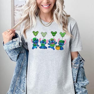 May include: A light gray t-shirt featuring a St. Patrick's Day design. The graphic shows four blue cartoon characters holding green balloons and wearing green hats. One character holds a pot of gold. The design is centered on the shirt.