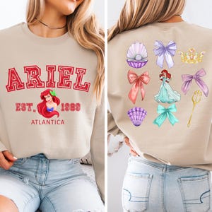 Disney Ariel sweatshirt in pastel blue with bold red varsity lettering on the front, reading 'Ariel Est. 1989 Atlantica.' The back design includes princess elements like seashells, bows, a golden trident, and an elegant illustration of