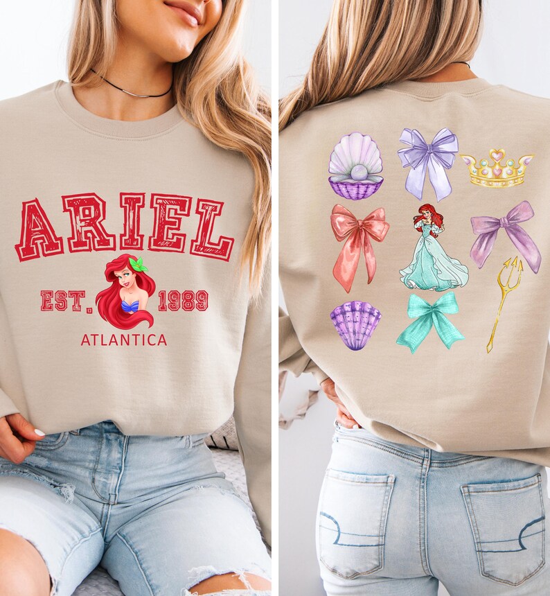 Disney Ariel sweatshirt in pastel blue with bold red varsity lettering on the front, reading 'Ariel Est. 1989 Atlantica.' The back design includes princess elements like seashells, bows, a golden trident, and an elegant illustration of