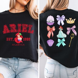 Disney Ariel sweatshirt in pastel blue with bold red varsity lettering on the front, reading 'Ariel Est. 1989 Atlantica.' The back design includes princess elements like seashells, bows, a golden trident, and an elegant illustration of