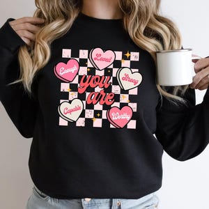May include: Black sweatshirt with a checkerboard design featuring pink hearts with the words "Enough", "Loved", "Strong", "Capable", and "Worthy". The words "you are" are in a cursive font. A white mug is held in the hand.