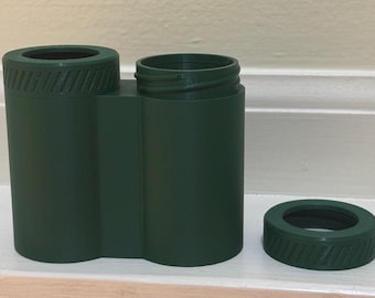 May include: A dark green, dual-cylinder container featuring a threaded top rim with diagonal grooves. One cylinder is capped with a matching dark green ring, while the other cylinder remains open, revealing the internal threading for a lid.