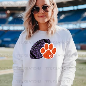 May include: White sweatshirt with a purple leopard print American football and an orange paw print with a heart. The background shows a stadium.