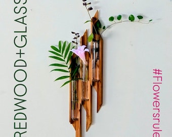 Pode incluir: Um vaso de parede de madeira e vidro com três tubos de vidro, contendo folhas verdes e uma flor rosa. O texto "REDWOOD+GLASS" está à esquerda e "#Flowersrule" à direita.