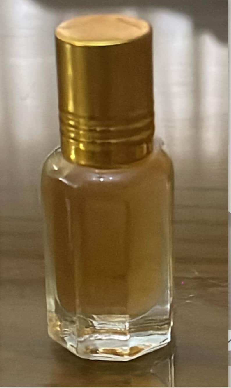 Pode incluir: Uma pequena garrafa de vidro transparente com uma tampa dourada. A garrafa cont&eacute;m um l&iacute;quido castanho claro. A tampa &eacute; cil&iacute;ndrica e tem um design canelado. A garrafa tem uma forma retangular com bordas chanfradas.