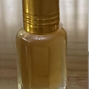 Könnte beinhalten: Kleine, klare Glasflasche mit goldfarbenem Deckel. Die Flasche enthält eine hellbraune Flüssigkeit. Der Deckel ist zylindrisch und hat ein geriffeltes Design. Die Flasche hat eine rechteckige Form mit abgeschrägten Kanten.