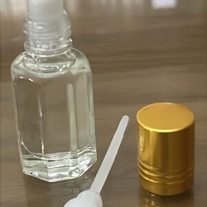 Puede incluir: Un frasco de perfume de vidrio transparente con una tapa de bola rodante blanca, un aplicador blanco y una tapa dorada. La botella est&aacute; vac&iacute;a y se encuentra sobre una superficie de madera. La tapa es dorada y tiene un dise&ntilde;o acanalado.