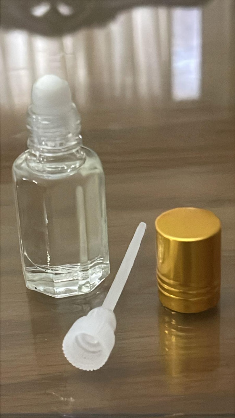 Puede incluir: Un frasco de perfume de vidrio transparente con una tapa de bola rodante blanca, un aplicador blanco y una tapa dorada. La botella est&aacute; vac&iacute;a y se encuentra sobre una superficie de madera. La tapa es dorada y tiene un dise&ntilde;o acanalado.