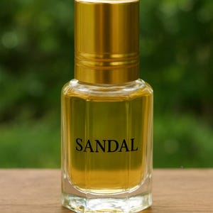 Pode incluir: Um frasco de perfume de vidro transparente com tampa dourada, preenchido com um l&iacute;quido &acirc;mbar claro. A palavra "SANDAL" est&aacute; impressa em preto na frente. O frasco est&aacute; sobre uma superf&iacute;cie de madeira.