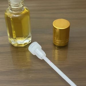 Pode incluir: Uma garrafa de vidro transparente cheia de l&iacute;quido dourado, uma tampa dourada e um aplicador branco com uma linha rosa. A garrafa tem uma tampa branca. Os itens est&atilde;o em uma superf&iacute;cie de madeira.
