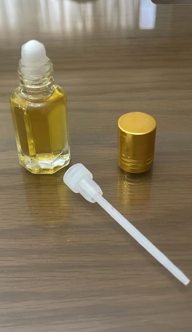 Pode incluir: Uma garrafa de vidro transparente cheia de l&iacute;quido dourado, uma tampa dourada e um aplicador branco com uma linha rosa. A garrafa tem uma tampa branca. Os itens est&atilde;o em uma superf&iacute;cie de madeira.