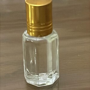 Puede incluir: Un peque&ntilde;o frasco de perfume de vidrio transparente con una tapa dorada. El frasco tiene forma rectangular con una base facetada. La tapa es cil&iacute;ndrica y tiene un acabado met&aacute;lico brillante. El frasco est&aacute; vac&iacute;o.