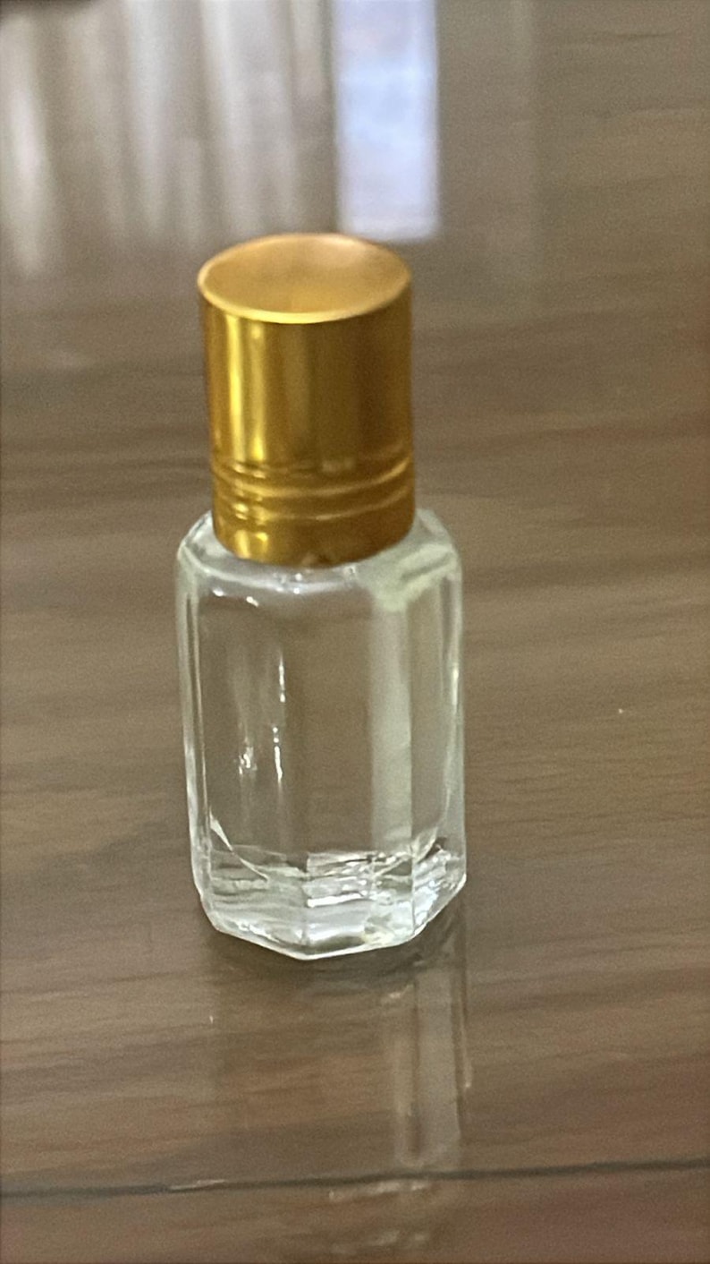 Puede incluir: Un peque&ntilde;o frasco de perfume de vidrio transparente con una tapa dorada. El frasco tiene forma rectangular con una base facetada. La tapa es cil&iacute;ndrica y tiene un acabado met&aacute;lico brillante. El frasco est&aacute; vac&iacute;o.