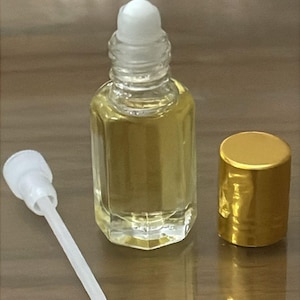 Puede incluir: Un frasco de perfume de vidrio transparente con una tapa de bola blanca y una tapa dorada. El frasco contiene un l&iacute;quido dorado. Un aplicador blanco est&aacute; al lado del frasco. Los art&iacute;culos est&aacute;n sobre una superficie marr&oacute;n.