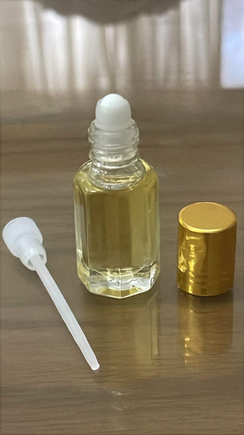 Puede incluir: Un frasco de perfume de vidrio transparente con una tapa de bola blanca y una tapa dorada. El frasco contiene un l&iacute;quido dorado. Un aplicador blanco est&aacute; al lado del frasco. Los art&iacute;culos est&aacute;n sobre una superficie marr&oacute;n.