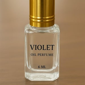 Puede incluir: Botella de perfume transparente y rectangular con tap&oacute;n dorado. La botella lleva la inscripci&oacute;n negra "VIOLET OIL PERFUME" y "6 ML". El fondo es borroso y de color neutro.