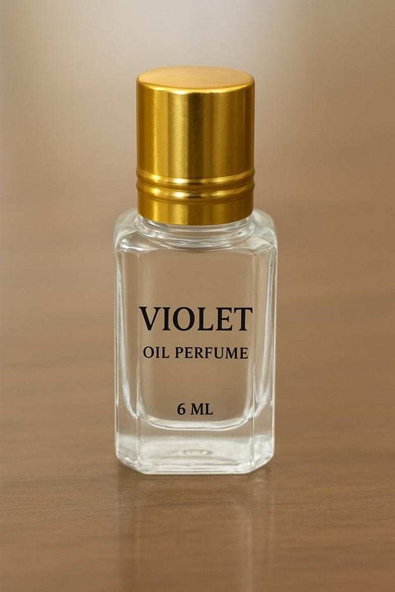 Puede incluir: Botella de perfume transparente y rectangular con tap&oacute;n dorado. La botella lleva la inscripci&oacute;n negra "VIOLET OIL PERFUME" y "6 ML". El fondo es borroso y de color neutro.
