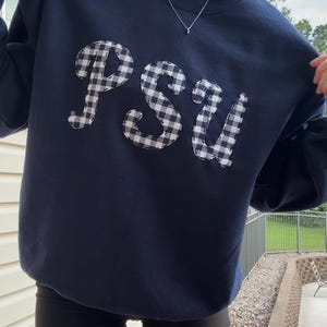 Peut inclure: Sweat-shirt bleu marine avec les lettres "PSU" appliquées dans un motif vichy blanc et noir. Le sweat-shirt est de couleur unie et a un col rond. L'image montre le sweat-shirt tenu.