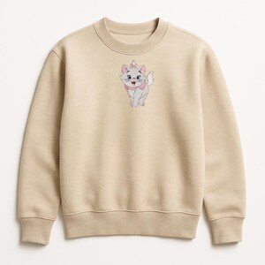 Puede incluir: Sudadera beige de cuello redondo con un dise&ntilde;o de gato de dibujos animados bordado. El gato tiene un lazo y una corona rosas, y es de color gris claro con detalles rosas. La sudadera tiene cuello, pu&ntilde;os y dobladillo de canal&eacute;.