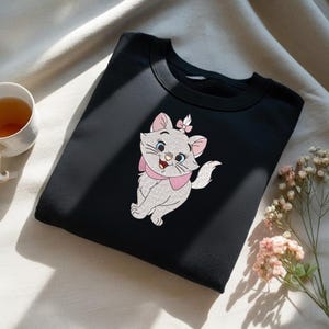 Puede incluir: Sudadera negra con un dise&ntilde;o de gato de dibujos animados. El gato es blanco con detalles rosas, un lazo y un collar. El gato tiene ojos azules y una expresi&oacute;n sonriente. La sudadera es de material suave.