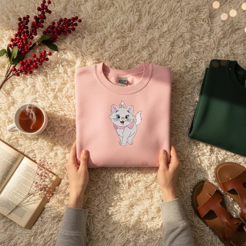 Puede incluir: Una sudadera rosa claro con un gato de dibujos animados bordado con un lazo y una corona rosas. La sudadera la sostiene una persona. Tambi&eacute;n se ven una sudadera verde oscuro, una taza de t&eacute;, un libro abierto y sandalias.