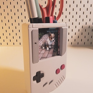 May include: A white desk organizer shaped like a retro handheld game console, with a photo of a corgi puppy. The organizer holds pens, scissors, and other office supplies. Red and pink accents are visible on the console.
