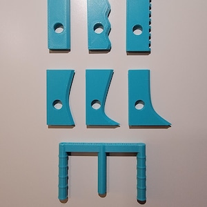 May include: A collection of seven turquoise 3D-printed tools, including various shapes of scrapers and a handle. Each tool has a hole for hanging or attachment. The tools are arranged on a light gray surface.