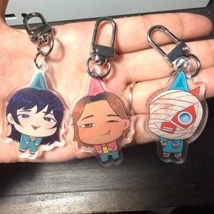 May include: Three clear acrylic keychains with cartoon character designs. Each keychain features a different character with unique facial expressions and outfits. The keychains have silver-colored metal clasps for easy attachment to bags or keys.