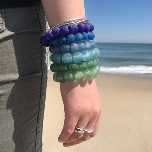 May include: A stack of colorful glass bead bracelets in shades of blue, green, and teal. The bracelets are worn on a person's wrist.