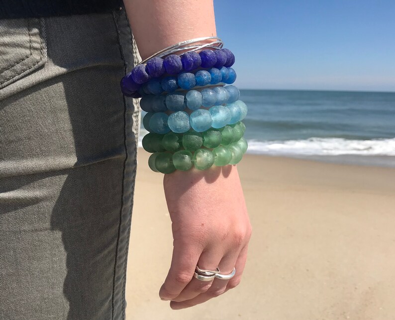May include: A stack of colorful glass bead bracelets in shades of blue, green, and teal. The bracelets are worn on a person's wrist.