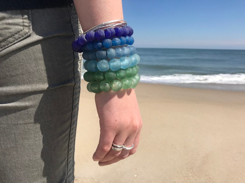 May include: A stack of colorful glass bead bracelets in shades of blue, green, and purple. The bracelets are worn on a person's wrist.