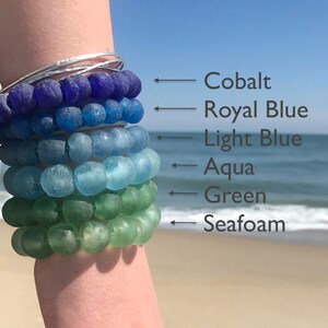 May include: A stack of six bracelets made of glass beads in shades of blue and green. The bracelets are labeled from top to bottom: Cobalt, Royal Blue, Light Blue, Aqua, Green, and Seafoam.