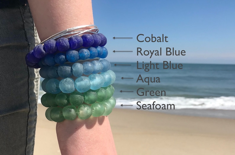 May include: A stack of six bracelets made of glass beads in shades of blue and green. The bracelets are labeled from top to bottom: Cobalt, Royal Blue, Light Blue, Aqua, Green, and Seafoam.