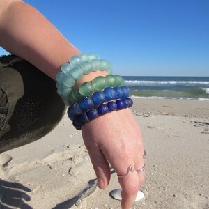 May include: A stack of four bracelets made of blue, green, and aqua glass beads. The bracelets are worn on a person's wrist against a sandy beach background.