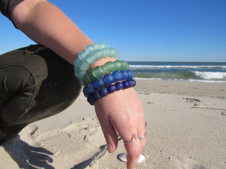 May include: A stack of four bracelets made of blue, green, and aqua glass beads. The bracelets are worn on a person's wrist against a sandy beach background.