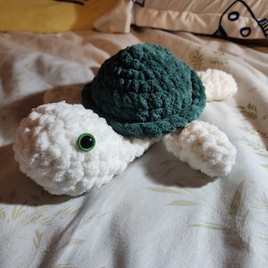 May include: A handmade crochet turtle toy. The turtle features a dark green shell, white head and limbs, and a black eye with a green border. The toy is made of soft, textured yarn.