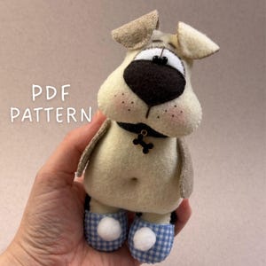 May include: A handmade plush dog with a black nose, floppy ears, and a bone-shaped tag. The dog wears blue and white checkered slippers and has the text "PDF PATTERN" in the upper left corner. The dog is cream-colored with brown accents.