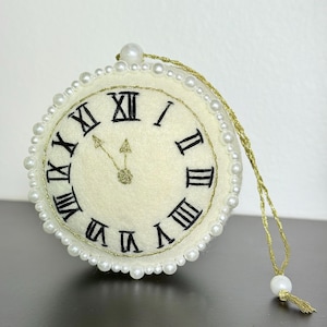 Op de afbeelding: Crèmekleurig vilten klokornament met zwarte Romeinse cijfers en goudkleurige wijzers. De klok is afgewerkt met imitatieparels en heeft een goudkleurig ophangkoord. De klok geeft ongeveer 12:05 uur aan.