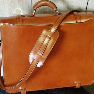 May include: A brown leather briefcase with a long shoulder strap and a top handle. The briefcase has a flap closure and a metal buckle.
