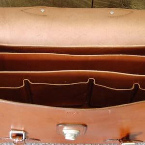 May include: A brown leather briefcase with multiple compartments and pockets. The briefcase has a flap closure with a metal buckle.