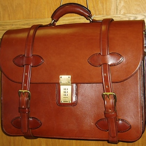 May include: A brown leather briefcase with a combination lock and a shoulder strap. The briefcase has a flap that closes with a buckle.