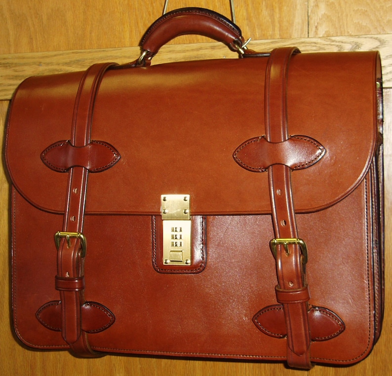 May include: A brown leather briefcase with a combination lock and a shoulder strap. The briefcase has a flap that closes with a buckle.